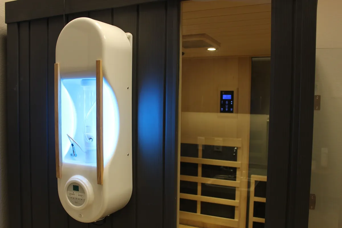 A wall-mounted halotherapy generator lit blue beside the glass door of the cedar infrared sauna.