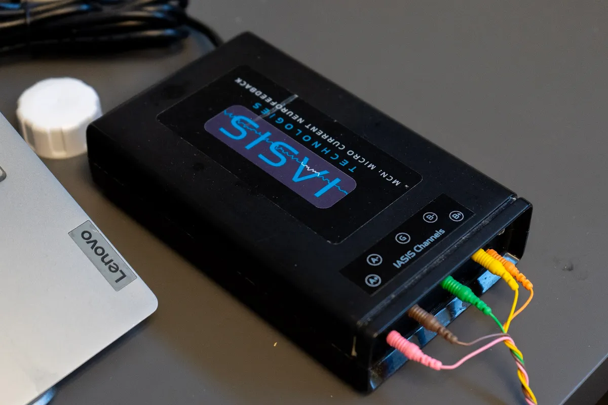 The small IASIS microcurrent neurofeedback unit on a desk, its coloured leads plugged into the side, beside a laptop.