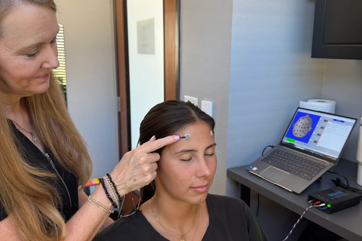 A practitioner placing a small round sensor on the forehead of a seated client with their eyes closed, a laptop showing a head map on the desk behind.