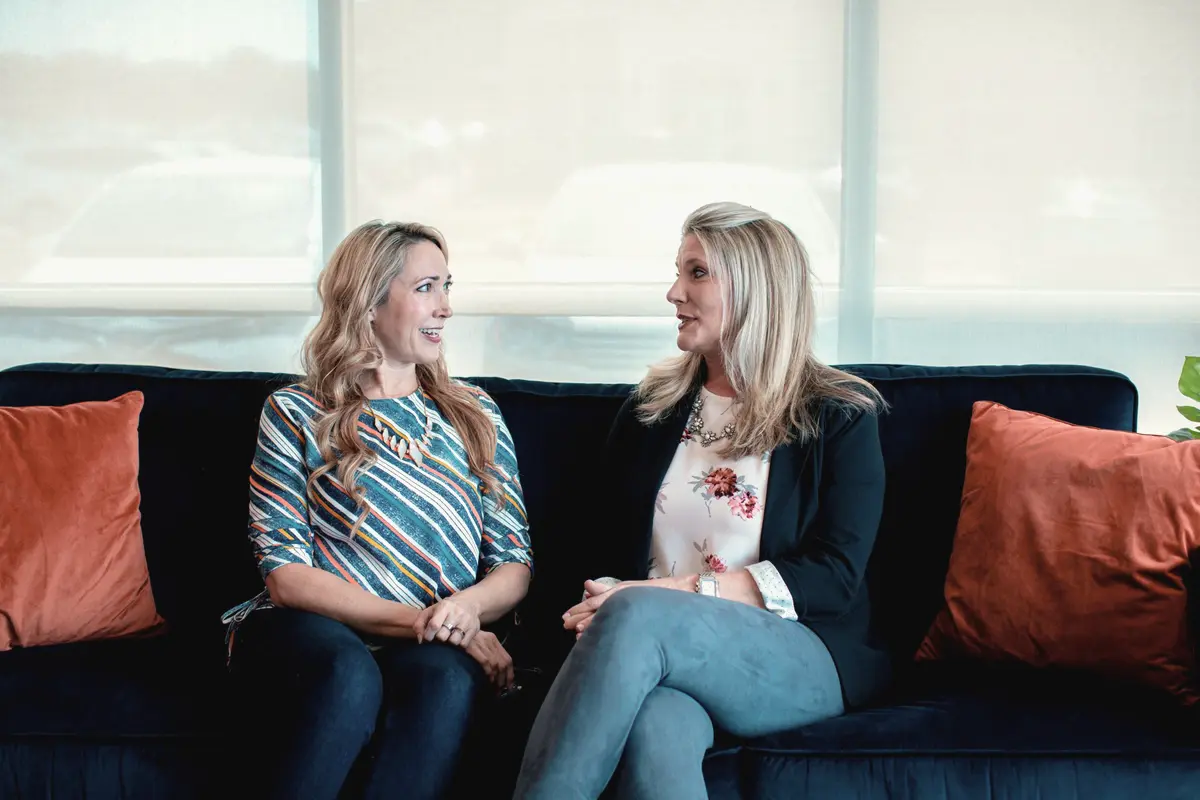 Two people sitting and talking on a dark sofa in front of a bright window, cushions either side.