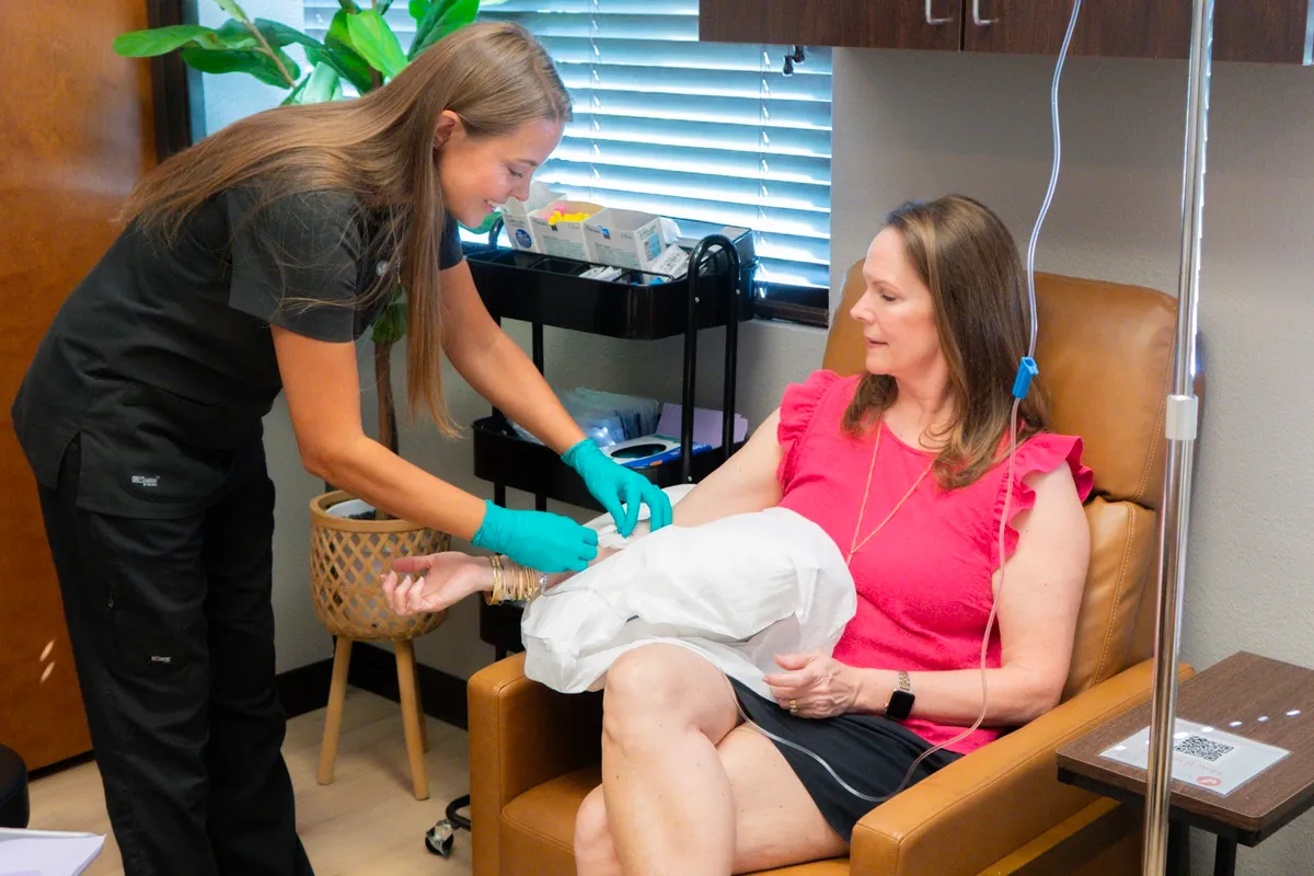 A practitioner in scrubs and gloves working at the arm of a client seated in a tan leather recliner, a drip stand and supply cart alongside.