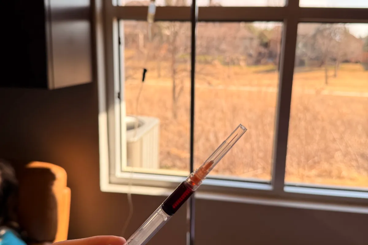 A blood-collection syringe held up at a window, with open fields visible outside.