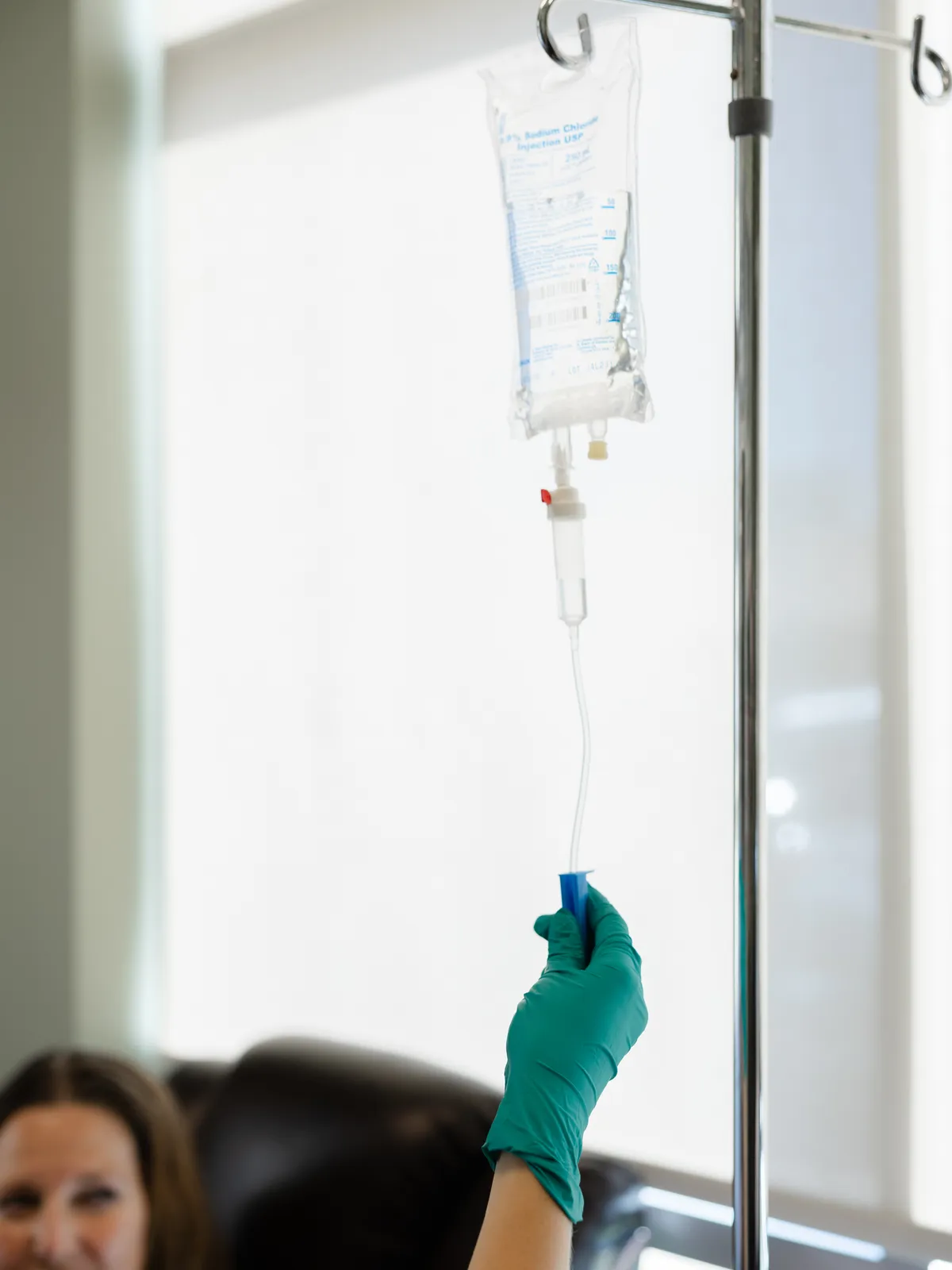 An IV bag on the pole, backlit by the window, a gloved hand on the line